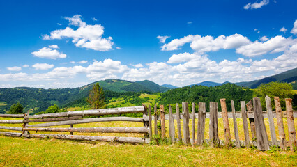 Fototapeta na wymiar wooden fence on the meadow. mountainous rural landscape of transcarpathia, ukraine in summer. carpathian countryside with forested rolling hill beneath a blue sky with white fluffy clouds
