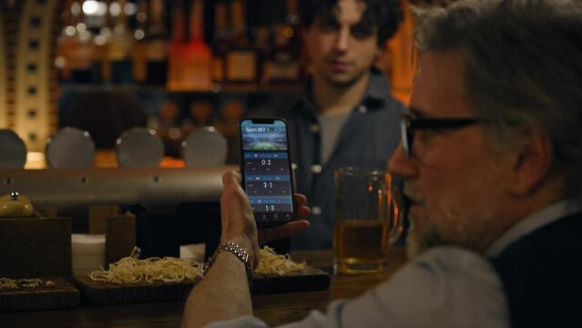 Mature sports fans check game score and bookmaker ratings in app using mobile phone, talk with bartender. Male friends bet on football match online sitting at bar counter in pub. Gambling concept.
