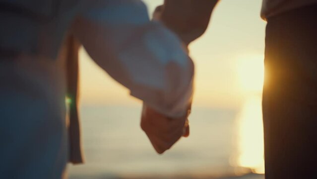 Hands of elderly people at sunset. Married old couple man woman holding hands and walking along the seashore together, back view. Long-marriage love, family support, together vacation concept.