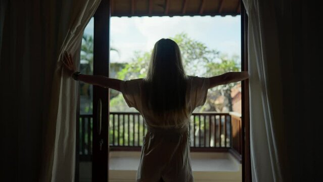Woman going out balcony, enjoying view. Young woman opens curtains and looks to the beautiful landscape sea from villa balcony, enjoying rest. Vacation summer morning, resort, hotel relax concept.