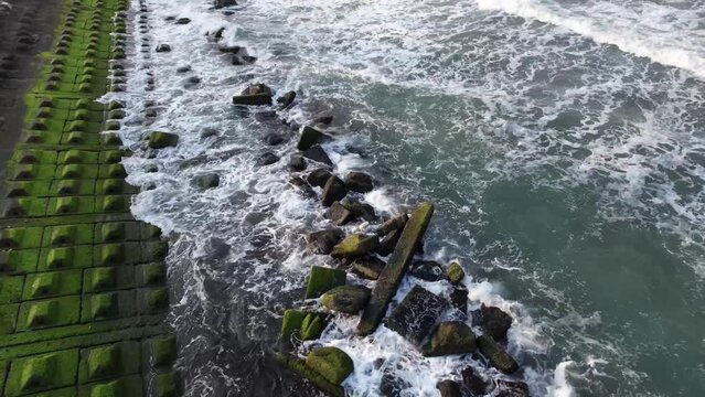 Aerial drone footage of foamy waves breaking on moss-covered tetrapods at a coastal sea defense, illustrating natures power and coastal engineering