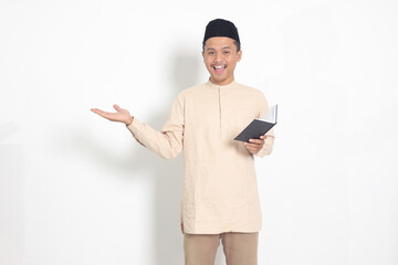Portrait of excited muslim man in koko shirt with peci reading a notebook. Happy Asian guy pointing...
