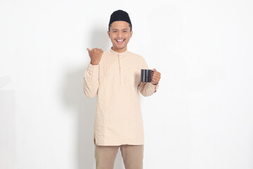 Portrait of attractive Asian muslim man in koko shirt with peci holding a mug, drinking water,...