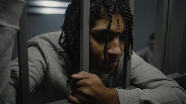 Close up shot of angry African American teenage prisoner with face tattoos standing in prison cell, holding metal bars. Young inmate serves imprisonment term for crime in jail. Youth detention center.