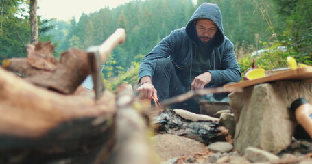 A hiker in a hood cooks food on a campfire near a tent in a camp.