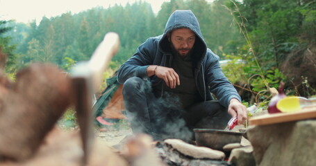 A hiker in a hood cooks food on a campfire near a tent in a camp.