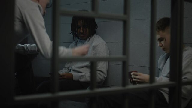 Three diverse teenagers play card games in prison cell. Young prisoners, criminals serve imprisonment term for crimes in jail. Youth detention center or correctional facility. View through metal bars.