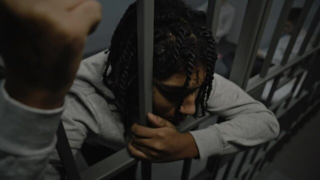 Upset African American teenage prisoner stands in prison cell in jail, holds metal bars. Young inmates play cards on bed in the background. Youth detention center or correctional facility. High angle.