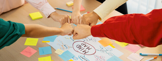 Young happy creative startup group join circle fist bump hands together surrounded by marketing strategy mind map and colorful sticky notes at meeting room. Unity and teamwork concept. Variegated. - obrazy, fototapety, plakaty