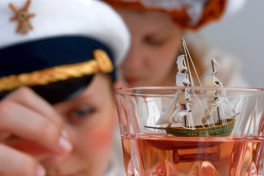 toy ship in a glass of rose, person wearing a sailor hat in the background