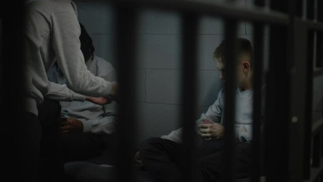 Multicultural young criminals play card games in prison cell. Teenage prisoners serve imprisonment term for crimes in jail. Youth detention center or correctional facility. View through metal bars.