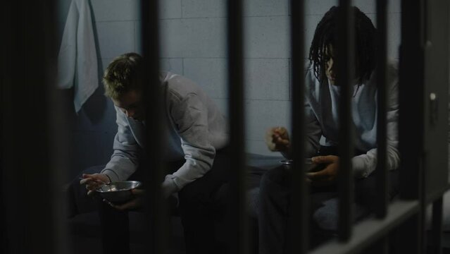 Two multiethnic teenage prisoners sit on bed in prison cell and eat food from iron bowls. Young criminals serve imprisonment term for crimes in jail. Youth detention center or correctional facility.