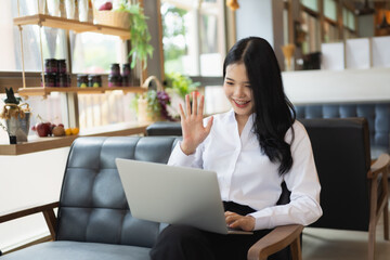 Businesswoman sitting on a couch and using a laptop. She is smiling and waving at the camera. Concept of warmth and friendliness, as the woman is enjoying her time on the couch while using her laptop