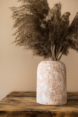 A clay vase with a bouquet of dried pompas grass on an old wooden table. Sharp shadows. Dark tones....