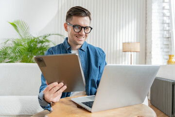 Joyful man with glasses multitasking with a tablet and laptop at a wooden table in a bright room.