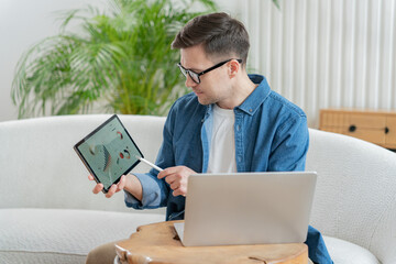 Man in glasses reviews financial data on tablet with stylus, with laptop on wooden table.