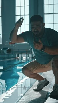 Medium vertical footage of Asian or Middle Eastern male coach crouching by pool side, cheering at sportsman in water, waving arm, looking at stopwatch, urging to speed up during training session