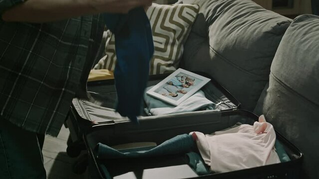 Lowkey cropped shot of unrecognizable woman going through domestic violence and abuse packing suitcase with clothes and picture of her family in frame while leaving dark cold apartment