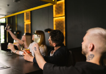 Group of friends takes selfie while raising glasses of drinks. Blonde lady takes pictures on smartphone sitting at bar counter