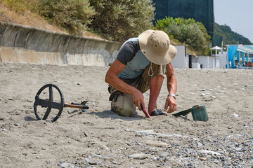 A man wearing shorts and a hat, with a metal detector, is searching for gold and coins on a sandy beach the daytime in summer.