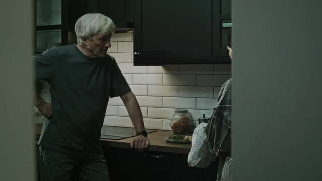 Medium lowkey shot of Caucasian adult abusive husband harassing and shouting at his terrified wife washing dishes at kitchen after dinner