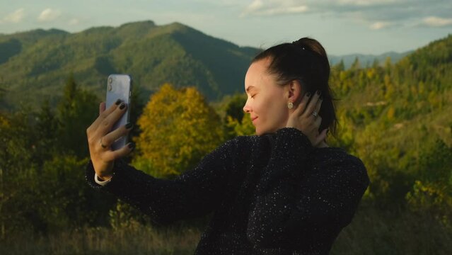 Young woman taking selfies using mobile phone standing on top of mountain peak. Happy girl making photos for social media on cellphone camera, scenic landscape view