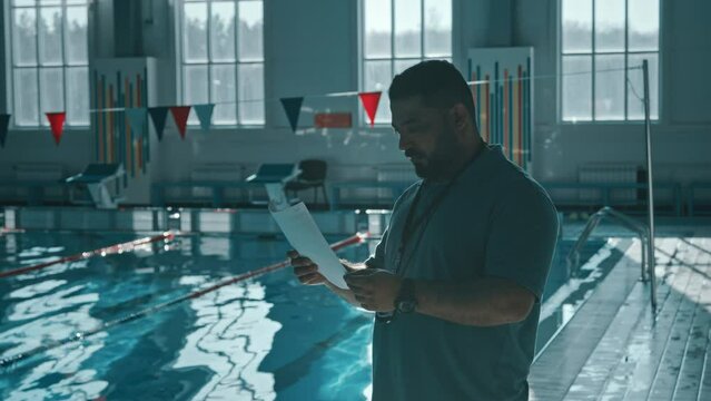 Medium shot with lens flare of middle-aged biracial male swimming coach standing on edge of pool before training session, looking through list of swimmers and their results