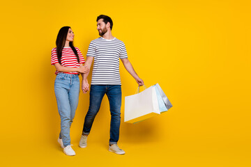 Full size photo of cheerful man look at girlfriend hold shoppings bags go to empty space isolated on vibrant yellow color background