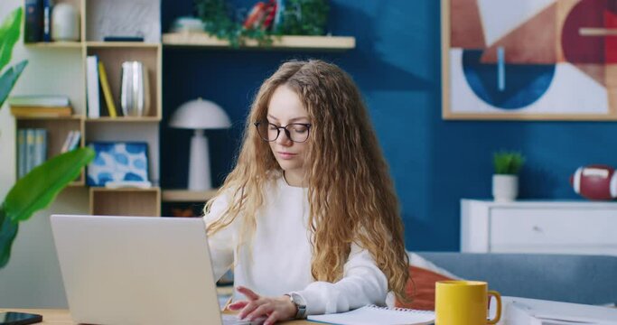 Red-haired businesswoman opening and typing on her laptop. Young professional woman writing important email to customer from home. Modern living room office. Remote work, freelancing.