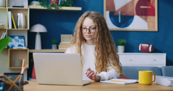 Smiling young woman chatting in internet on laptop indoor home background. Cheerful girl sitting at table desk using notebook pc communicating with friends slow motion. Online connection