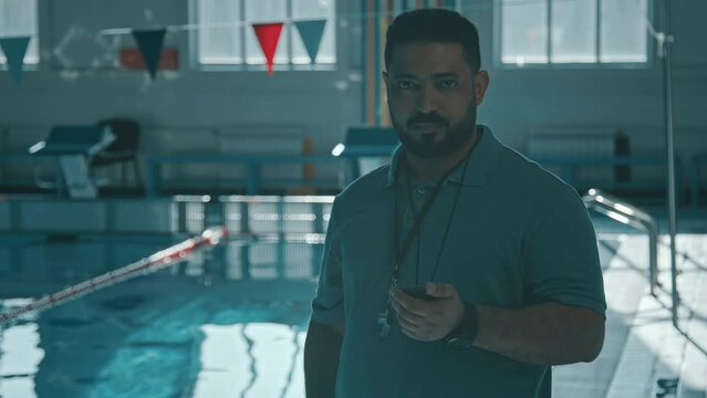 Medium shot with lens flare of Middle Eastern male swim instructor with whistle standing in pool, checking stopwatch, looking at camera, while supervising training session for professional athletes