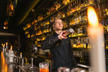 Professional barkeeper mixes cocktail ingredients using shaker by bar counter. Barman makes elite beverages for guests of popular pub