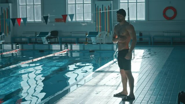 Full shot of young muscular Caucasian male swimmer standing by pool, doing warmup, rotating shoulders, flexing, swinging arms to prepare for practice, coach walking up, both discussing training goals