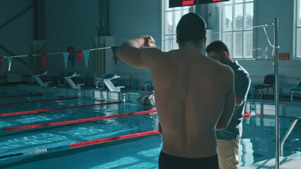 Medium rear follow shot of male swimmer walking out of changing room, rotating arms, warming up wrists, greeting coach, who is showing practice plan on smartphone and setting goals for workout