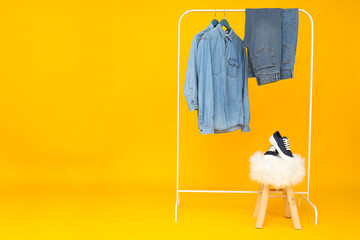 Denim pants and shirts on hangers, shoes on stool on yellow background, space for text