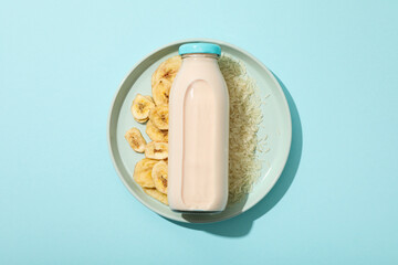 Bottle with milk on plate with rice and sliced bananas on blue background, top view