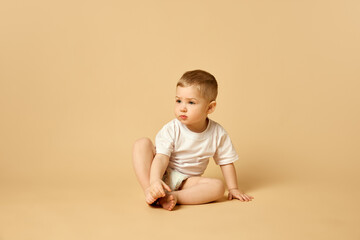 Cute little baby, boy, child in diaper and t-shirt calmly sitting on bed isolated over beige studio background. Concept of childhood and family, care, parenthood, infancy and heath
