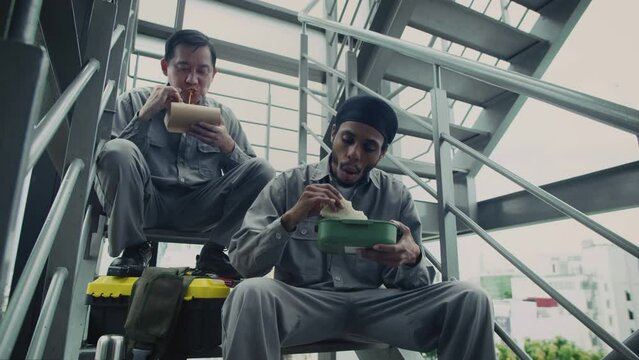 Low angle shot of foreman and builder in gray uniform eating lunch while resting on stairs