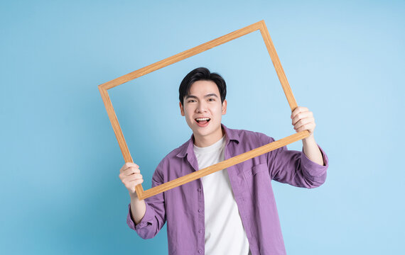 Young Ásian man holding photo frame on blue background
