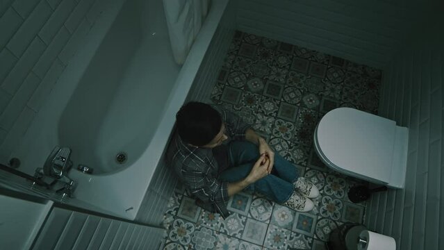 Top view lowkey shot of sad Caucasian adult woman sitting on cold tile in dark bathroom alone