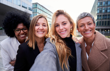 Cheerful selfie of four multiracial businesswomen using phone to take self-photos together outside workplace looking at camera smiling.