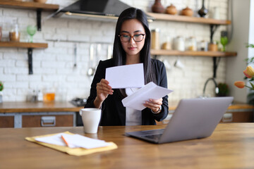 A young businesswoman sits at home, smiling while reading paperwork on her laptop.