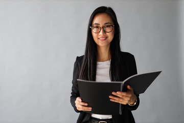 A young Asian businesswoman stands confidently, holding a folder, smiling in her professional grey office.