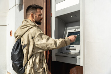 Young fashionable hipster backpacker traveler man using credit card to withdraw money from ATM...