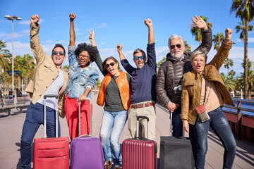 Group of middle aged excited friends posing for the camera happily celebrating their holidays. Diverse people with arms raised and their luggage in the street on a sunny winter day