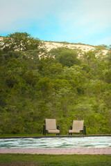Fototapeta na wymiar Two comfortable lounge chairs sit empty, inviting relaxation by the side of a tranquil pool, set against a backdrop of dense green foliage and gently sloping hills. The setting sun casts a warm, soft 