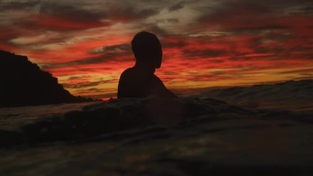 Medium shot of silhouette of man wading in water with board under red fire sunset sky