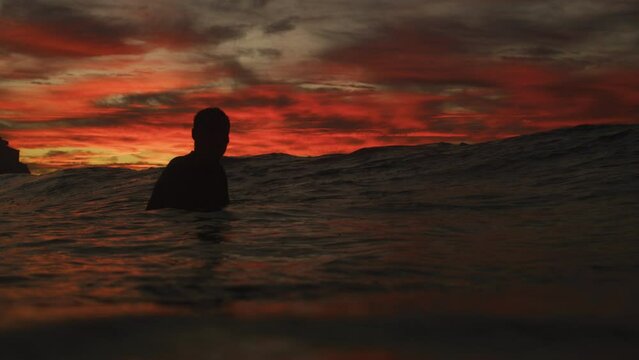 Man turns head waiting in shallow water at sunset, view from ocean surface with red sky