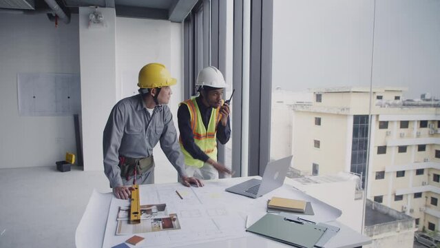 Wide shot of diverse male engineers checking workplan standing near table with blueprints and using walkie-talkie