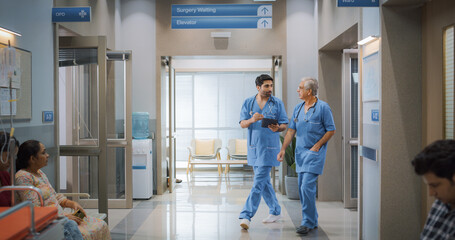 Portrait of Two Indian Male Doctors Walking in Hospital Corridor and Talking while Using a Digital...
